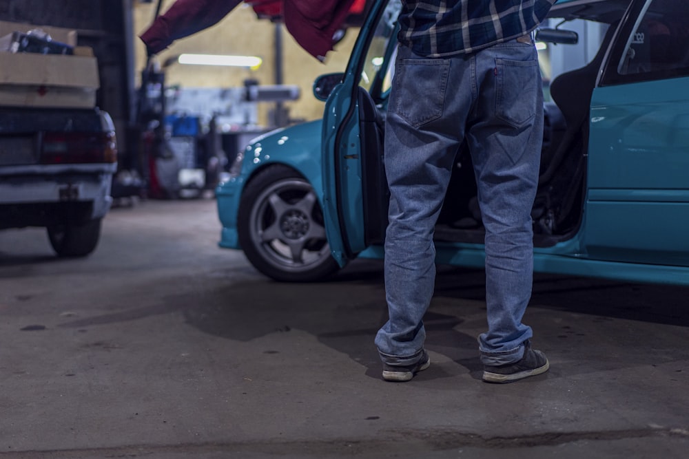 man in blue denim jeans and black leather boots standing beside blue car during daytime