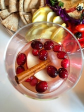 A glass of beverage is filled with cranberries, apple slices, and a cinnamon stick. Surrounding the glass is a platter with sliced pita bread and an assortment of vegetables including tomatoes, cucumber slices, and purple cabbage.