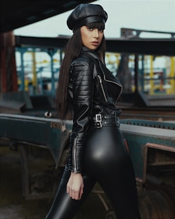 A person wearing a black leather outfit with a biker jacket and a beret poses confidently in an industrial setting with metal structures and muted colors in the background.