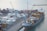 A busy shipping port with numerous stacked cargo containers is shown. Several cranes are actively loading and unloading the containers onto a large cargo ship docked at the port. The water nearby appears calm, and there are various vehicles and equipment scattered around the area, indicating a high level of activity.