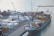 A busy shipping port with numerous stacked cargo containers is shown. Several cranes are actively loading and unloading the containers onto a large cargo ship docked at the port. The water nearby appears calm, and there are various vehicles and equipment scattered around the area, indicating a high level of activity.