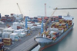 A busy shipping port with numerous stacked cargo containers is shown. Several cranes are actively loading and unloading the containers onto a large cargo ship docked at the port. The water nearby appears calm, and there are various vehicles and equipment scattered around the area, indicating a high level of activity.