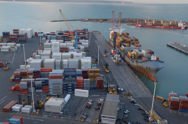 A busy port scene features numerous stacked shipping containers of various colors and labels, such as Maersk and Hamburg-Süd. A large cargo ship is docked alongside a pier, loaded with more shipping containers. Cranes and industrial machinery are actively engaged in loading or unloading the containers. The sea provides a calm backdrop, while the port infrastructure includes paved roads and parking areas with vehicles scattered around.