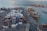A busy port scene features numerous stacked shipping containers of various colors and labels, such as Maersk and Hamburg-Süd. A large cargo ship is docked alongside a pier, loaded with more shipping containers. Cranes and industrial machinery are actively engaged in loading or unloading the containers. The sea provides a calm backdrop, while the port infrastructure includes paved roads and parking areas with vehicles scattered around.