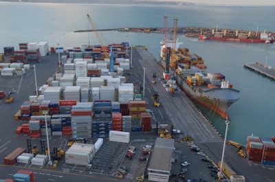 A busy port scene features numerous stacked shipping containers of various colors and labels, such as Maersk and Hamburg-Süd. A large cargo ship is docked alongside a pier, loaded with more shipping containers. Cranes and industrial machinery are actively engaged in loading or unloading the containers. The sea provides a calm backdrop, while the port infrastructure includes paved roads and parking areas with vehicles scattered around.