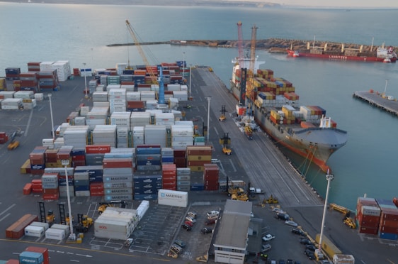 A busy port scene features numerous stacked shipping containers of various colors and labels, such as Maersk and Hamburg-Süd. A large cargo ship is docked alongside a pier, loaded with more shipping containers. Cranes and industrial machinery are actively engaged in loading or unloading the containers. The sea provides a calm backdrop, while the port infrastructure includes paved roads and parking areas with vehicles scattered around.