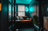 A cozy room with teal walls and a brown sofa adorned with cushions, one of which has 'Internet Famous' written on it. A neon light in the shape of a cursor arrow hangs on the wall above the sofa. The room is lit by natural light coming through the blinds on the window, and there's a green potted plant in the corner.