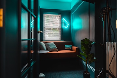 A cozy room with teal walls and a brown sofa adorned with cushions, one of which has 'Internet Famous' written on it. A neon light in the shape of a cursor arrow hangs on the wall above the sofa. The room is lit by natural light coming through the blinds on the window, and there's a green potted plant in the corner.