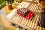 A neatly folded stack of colorful blankets rests on a wooden surface next to a woven basket. The surface is made of light wood planks supported by concrete blocks, and it sits on a textured beige mat. A potted fern plant with lush green leaves is seen in the background, casting soft shadows.