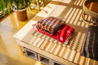 A neatly folded stack of colorful blankets rests on a wooden surface next to a woven basket. The surface is made of light wood planks supported by concrete blocks, and it sits on a textured beige mat. A potted fern plant with lush green leaves is seen in the background, casting soft shadows.