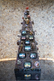 A tall, pyramid-like sculpture composed of multiple televisions stacked in a tiered structure. The screens display various images, adding a dynamic element to the piece. The structure is adorned with colorful wires and diverse decorative items, including masks and figurines. It stands in front of a textured stone wall, reflecting on the polished floor beneath.
