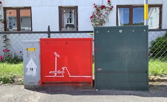 Three utility boxes are positioned in front of a residential building with a white exterior and brown window frames. The box on the left is gray with minimal features, the middle box is red and features graffiti art depicting a figure interacting with a camera on a stand, and the box on the right is green with no discernible markings. Behind the boxes, there is a chain-link fence with red flowers climbing up its surface, adding a touch of nature to the urban setting.