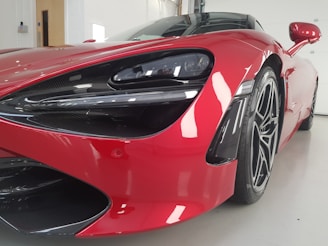 Close-up view of a luxurious red sports car with sleek design and aerodynamic features, showcased indoors in a well-lit space. The image highlights the car's front with a focus on the headlight and intricate detailing around the wheel.