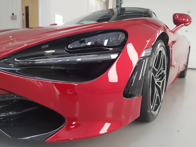 Close-up view of a luxurious red sports car with sleek design and aerodynamic features, showcased indoors in a well-lit space. The image highlights the car's front with a focus on the headlight and intricate detailing around the wheel.