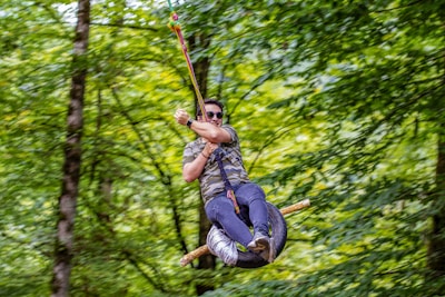 A person wearing sunglasses and a camouflage shirt is joyfully swinging on a zip line made of a tire in a lush green forest.