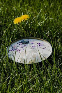 A round white candle decorated with delicate purple flower petals and a small decorative item in the center is placed on lush green grass. A yellow dandelion flower stands nearby, adding a contrasting pop of color to the scene.