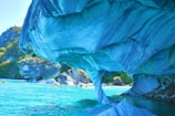 A stunning natural formation of marble caves with swirling blue and turquoise hues arches over crystal-clear waters. In the background, greenery is visible on rocky cliffs, while small kayaks with paddlers navigate the serene environment.