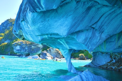 A stunning natural formation of marble caves with swirling blue and turquoise hues arches over crystal-clear waters. In the background, greenery is visible on rocky cliffs, while small kayaks with paddlers navigate the serene environment.
