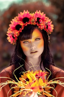 A woman with dark hair and striking makeup is wearing a vibrant headpiece made of large, bright sunflowers. Her face has a warm, golden-orange hue matching her makeup, with intricate detailing around the eyes and lips. She holds a bouquet of similarly colored flowers, and the background is softly blurred with earthy tones.