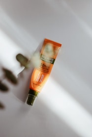 A tube of sunscreen labeled 'Solaire Peau Parfaite' with SPF 50+ UVA protection, placed on a light surface. The tube has an orange and green color scheme, and there are soft shadows and blurred leaves in the foreground, creating a natural setting.