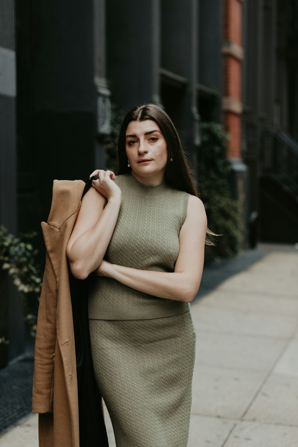 woman in gray tank top and brown coat standing on sidewalk during daytime