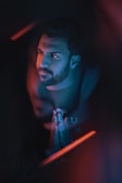 A bearded man with hands clasped in a thoughtful pose is surrounded by a blend of blue and red lighting, creating a reflective and contemplative atmosphere. The focus on the face is sharp while other parts are blurred, enhancing the mood of introspection.