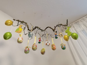 A decorative arrangement of multicolored Easter eggs and bunny cutouts hang from a twisted branch. The branch is suspended from the ceiling by thin, barely visible strings. The eggs are adorned with floral and Easter-themed patterns and ribbon loops. The background features a plain white wall and a partially visible curtain.