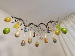 A decorative arrangement of multicolored Easter eggs and bunny cutouts hang from a twisted branch. The branch is suspended from the ceiling by thin, barely visible strings. The eggs are adorned with floral and Easter-themed patterns and ribbon loops. The background features a plain white wall and a partially visible curtain.