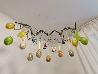 A decorative arrangement of multicolored Easter eggs and bunny cutouts hang from a twisted branch. The branch is suspended from the ceiling by thin, barely visible strings. The eggs are adorned with floral and Easter-themed patterns and ribbon loops. The background features a plain white wall and a partially visible curtain.