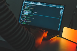 A shadowy figure typing code on a dimly lit laptop in a dark room.