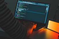 Close-up of hands typing code on a laptop with dark ambient lighting.
