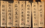 Several aged paper menus with Chinese calligraphy are displayed, each listing different dishes and their prices in yuan. The menus are attached to a wooden surface with nails.