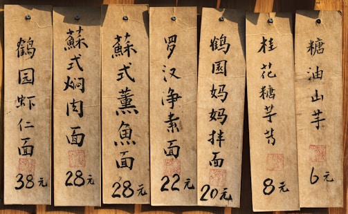 Several aged paper menus with Chinese calligraphy are displayed, each listing different dishes and their prices in yuan. The menus are attached to a wooden surface with nails.