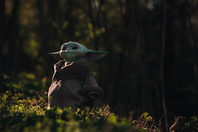 A small, green alien-like creature with large ears stands in a forest setting, wearing a brown robe. Soft light filters through the trees, casting a serene and mystical atmosphere.