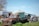 An old, rusted truck is parked in a grassy area, surrounded by various farm equipment and vehicles. The background features a large metal tank, a tractor, and a warehouse building. Several pieces of industrial machinery and pipes are scattered around the scene.