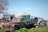 An old, rusted truck is parked in a grassy area, surrounded by various farm equipment and vehicles. The background features a large metal tank, a tractor, and a warehouse building. Several pieces of industrial machinery and pipes are scattered around the scene.