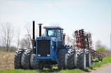 A large blue tractor with eight wheels is positioned in a rural agricultural setting. The machinery is robust, featuring a cab in the center and an attached red farming implement behind it. The background consists of a partly cloudy sky and bare, skeletal trees, suggesting an early spring or late fall season.