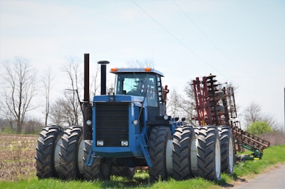 A large blue tractor with eight wheels is positioned in a rural agricultural setting. The machinery is robust, featuring a cab in the center and an attached red farming implement behind it. The background consists of a partly cloudy sky and bare, skeletal trees, suggesting an early spring or late fall season.