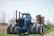 A large blue tractor with eight wheels is positioned in a rural agricultural setting. The machinery is robust, featuring a cab in the center and an attached red farming implement behind it. The background consists of a partly cloudy sky and bare, skeletal trees, suggesting an early spring or late fall season.