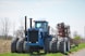 A large blue tractor with eight wheels is positioned in a rural agricultural setting. The machinery is robust, featuring a cab in the center and an attached red farming implement behind it. The background consists of a partly cloudy sky and bare, skeletal trees, suggesting an early spring or late fall season.