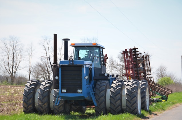 A large blue tractor with eight wheels is positioned in a rural agricultural setting. The machinery is robust, featuring a cab in the center and an attached red farming implement behind it. The background consists of a partly cloudy sky and bare, skeletal trees, suggesting an early spring or late fall season.