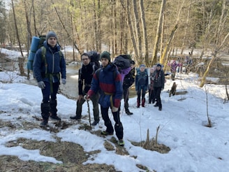 A group of hikers stands in a snowy woodland area. They are equipped with backpacks and outdoor gear, suggesting they are on a trekking trip. The background features a leafless forest with patches of snow on the ground, and a small stream flows through the scene.