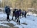 A group of hikers stands in a snowy woodland area. They are equipped with backpacks and outdoor gear, suggesting they are on a trekking trip. The background features a leafless forest with patches of snow on the ground, and a small stream flows through the scene.