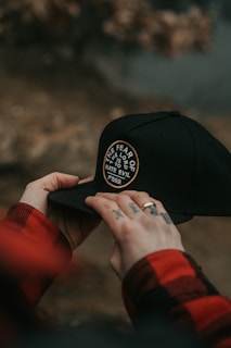 Hands holding a black cap with a circular patch that reads 'THE FEAR OF THE LORD IS TO HATE EVIL' and 'FSCO'. The person has tattoos on their fingers and is wearing a red and black checkered shirt.
