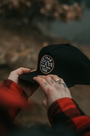 Hands holding a black cap with a circular patch that reads 'THE FEAR OF THE LORD IS TO HATE EVIL' and 'FSCO'. The person has tattoos on their fingers and is wearing a red and black checkered shirt.