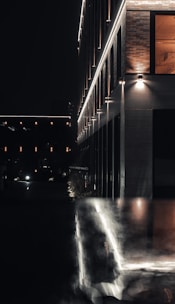 A modern building illuminated by exterior lighting at night, with reflective surfaces capturing the glow. The architecture features clean lines and a combination of brick and metallic panels. The scene is dark with minimal ambient lighting, creating a moody urban atmosphere.