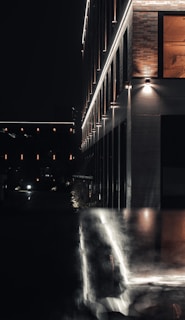 A modern building illuminated by exterior lighting at night, with reflective surfaces capturing the glow. The architecture features clean lines and a combination of brick and metallic panels. The scene is dark with minimal ambient lighting, creating a moody urban atmosphere.