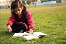 A young girl reading a book under a tree with a volunteer guiding her.