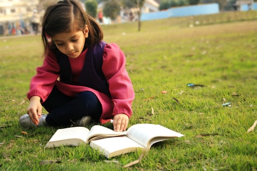 A young girl reading a book under a tree, surrounded by nature and sunlight.
