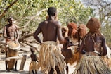 A group of people wearing traditional attire are gathered in an outdoor setting with a vibrant natural backdrop. The individuals are dressed in skirts made from natural materials and have unique hairstyles. One person is playing a large drum while others appear to be dancing or engaging in a ritual.